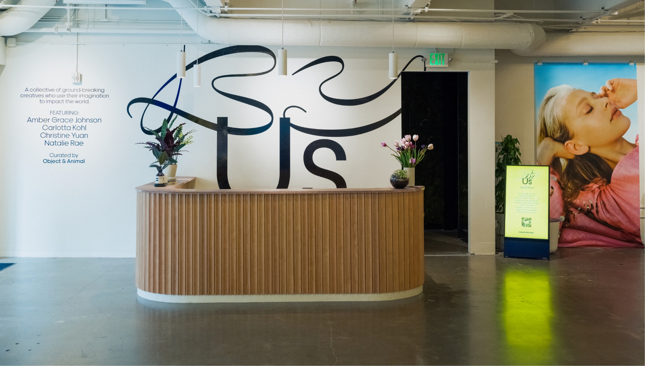 Front desk of art gallery with By Us large vinyl lettering on wall behind desk