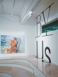 Rear corner of gallery showing stadium seating, video still print of woman floating in water with head in clear covering. Words By Us in vinyl on wall