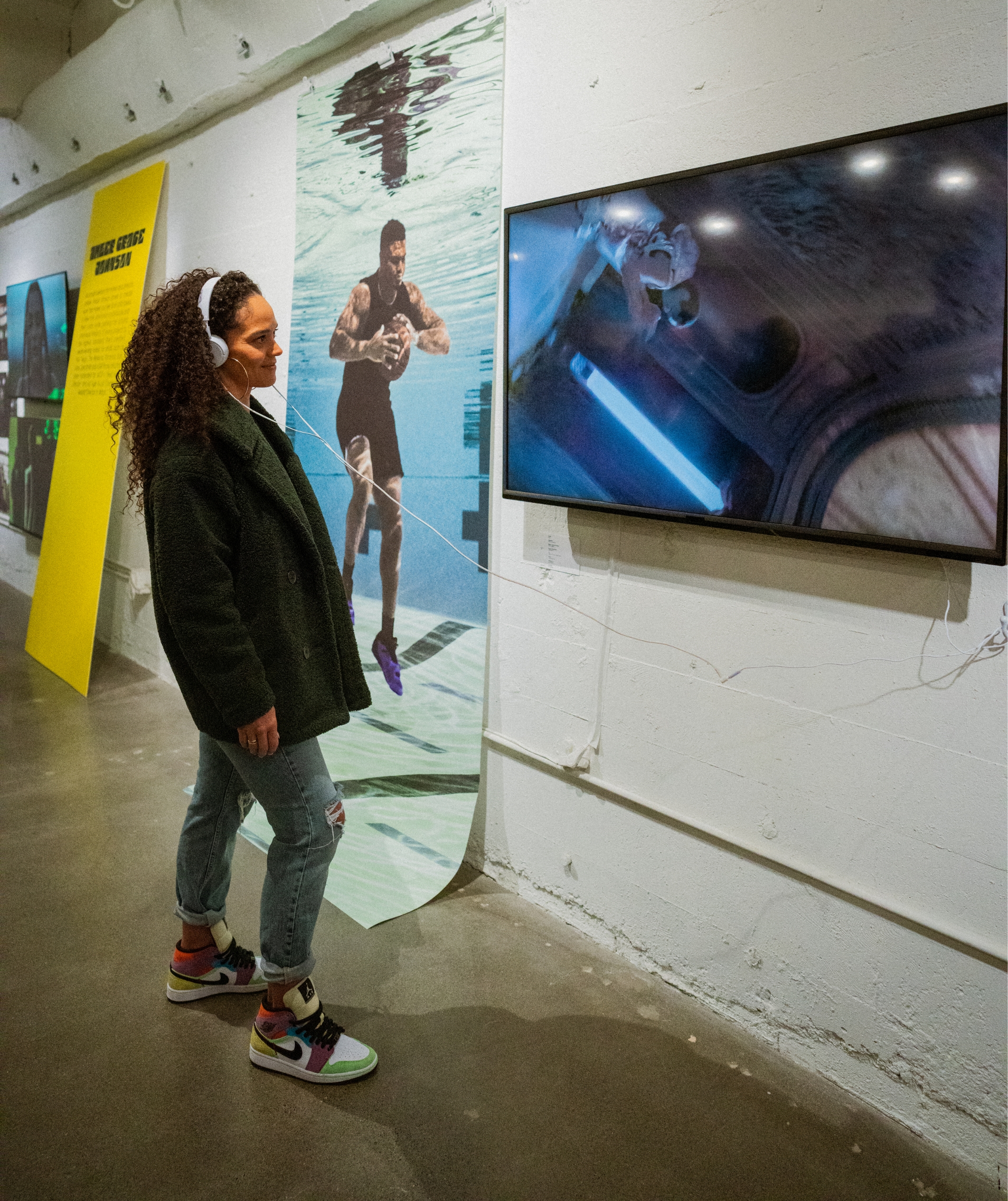 Woman in gallery watching TV mounted on wall with headphones on that are connected to the TV