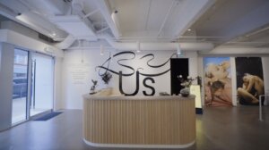 Front desk of art gallery with By Us large vinyl lettering on wall behind desk