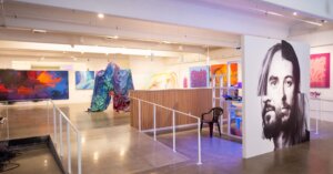 Sentiments Art show in Industry One gallery. Several paintings line 3 walls. A montage of two artists' faces is in center of gallery. Video screens on left wall. Tarp sculpture in foreground.