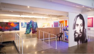 View of full gallery show from entrance. Painted tarp in back, large face montage, painted retro artifacts are prominent with walls lined with spray painted canvases