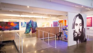 Sentiments Art show in Industry One gallery. Several paintings line 3 walls. A montage of two artists' faces is in center of gallery. Video screens on left wall. Tarp sculpture in foreground.