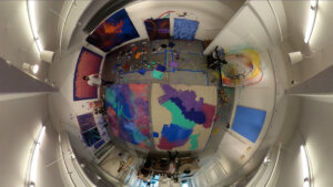 overhead fisheye lense view of Artist painting with paint buckets and paint trays on floor. Painted drop cloths cover the floor.