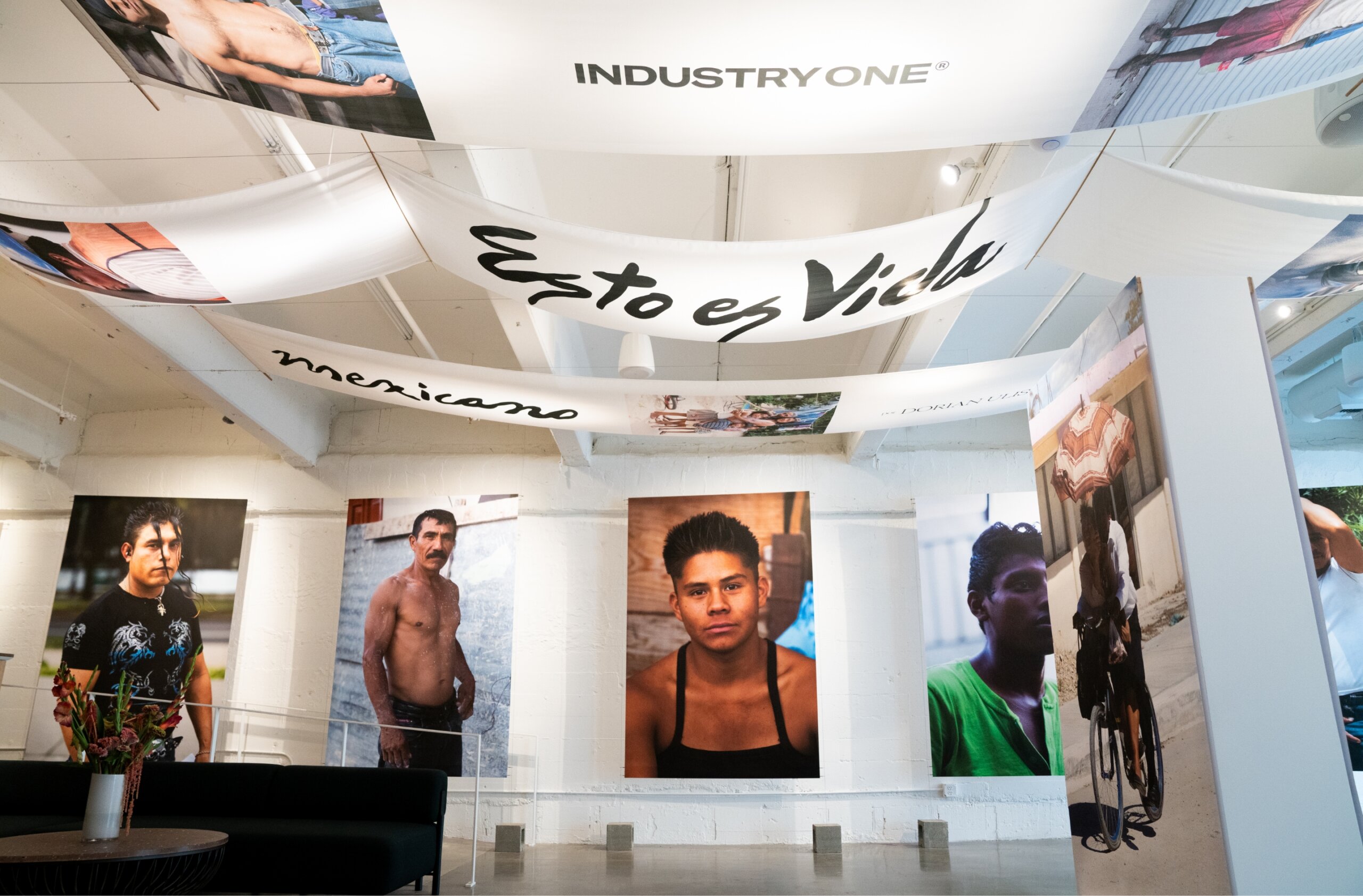 Art Gallery with fabric banners on ceiling reading INDUSTRY ONE, Esto es Vida and mexicana. Large portrait photos are hung on the wall