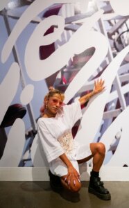 person in sunglasses and long white shirt crouches in front of gallery wall