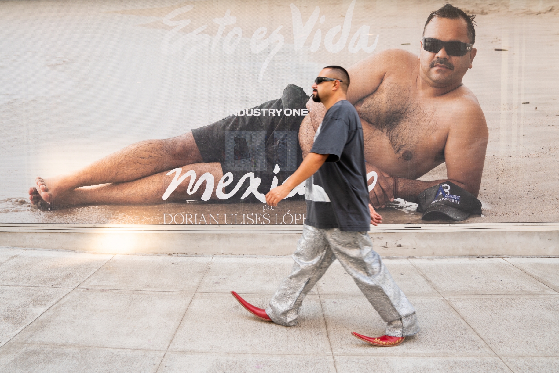 Dorian Lopez wearing Botas picudas walks on sidewalk in front of his Gallery show window. The window shows a Mexican man laying on beach with text reading esto es vida Industry One Mexicano