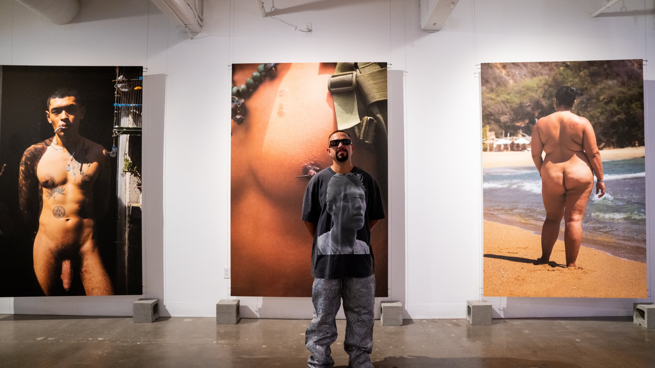 Dorian standing in front of 3 of his prints. One print is a front of naked man. Second is close up of man's nipple. Third is rear view of naked woman on beach.