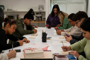 Several students sit around table sketching and carving wood blocks for prints
