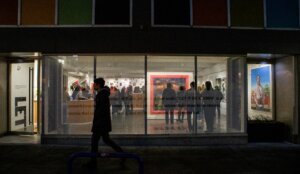 night time phonto of Industry One art Gallery front window. A person walks by on sidewalk and the inside of gallery is lit up showing many people and art pieces inside the gallery