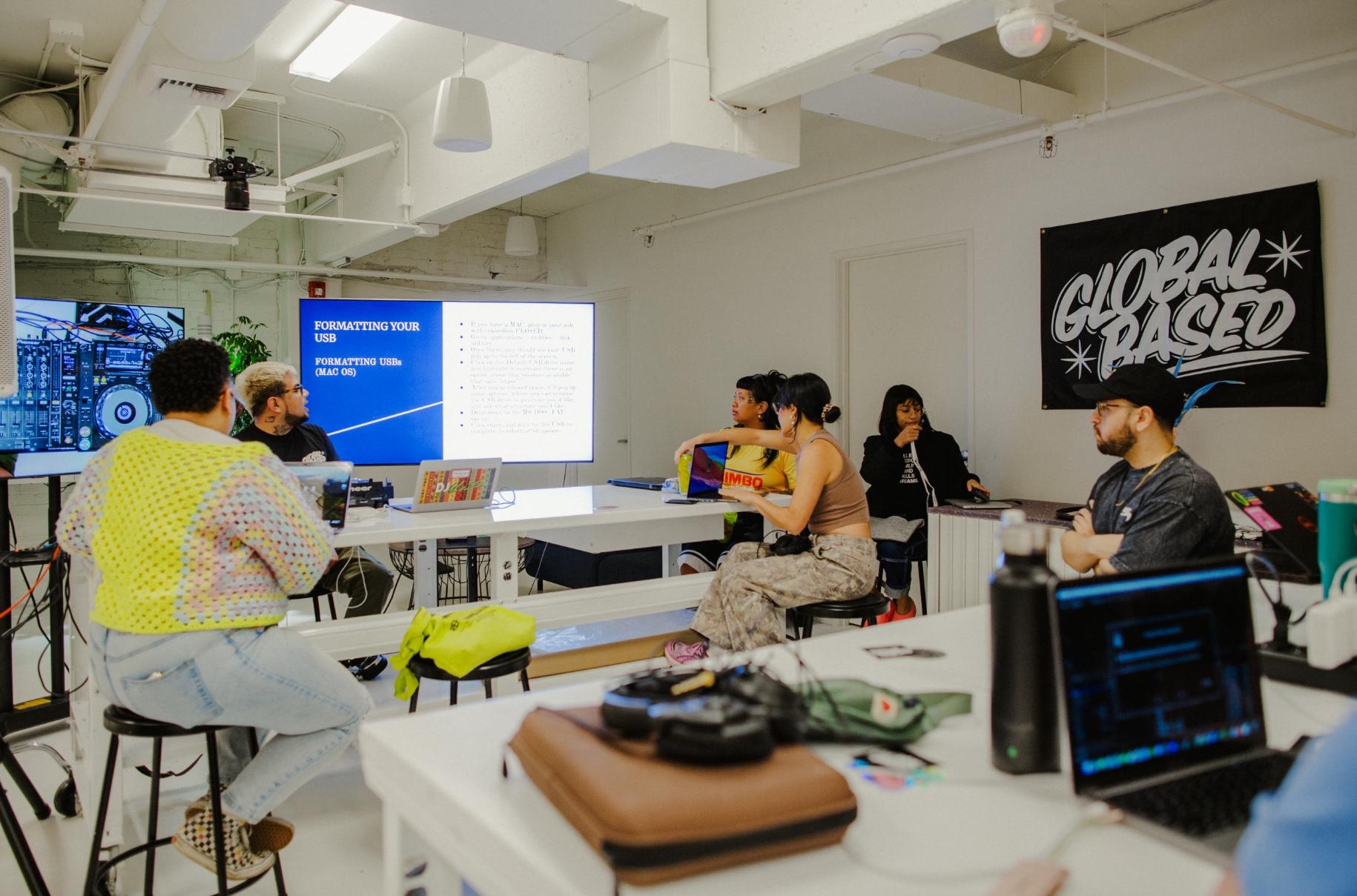 A group of people sitting in a modern classroom, engaging in a discussion while a presentation is displayed on two screens at the front of the room. A "Global Based" poster is on the wall.
