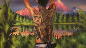 A person with extensive back tattoos, including a ram and a person's face, stands in front of a scenic mountain landscape at sunset with their arms raised and a gun tucked into their waistband.