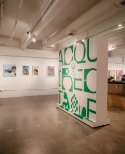 Art gallery interior with large green and white abstract letter display in the foreground. Framed artwork hangs on white walls. Track lighting illuminates the space.