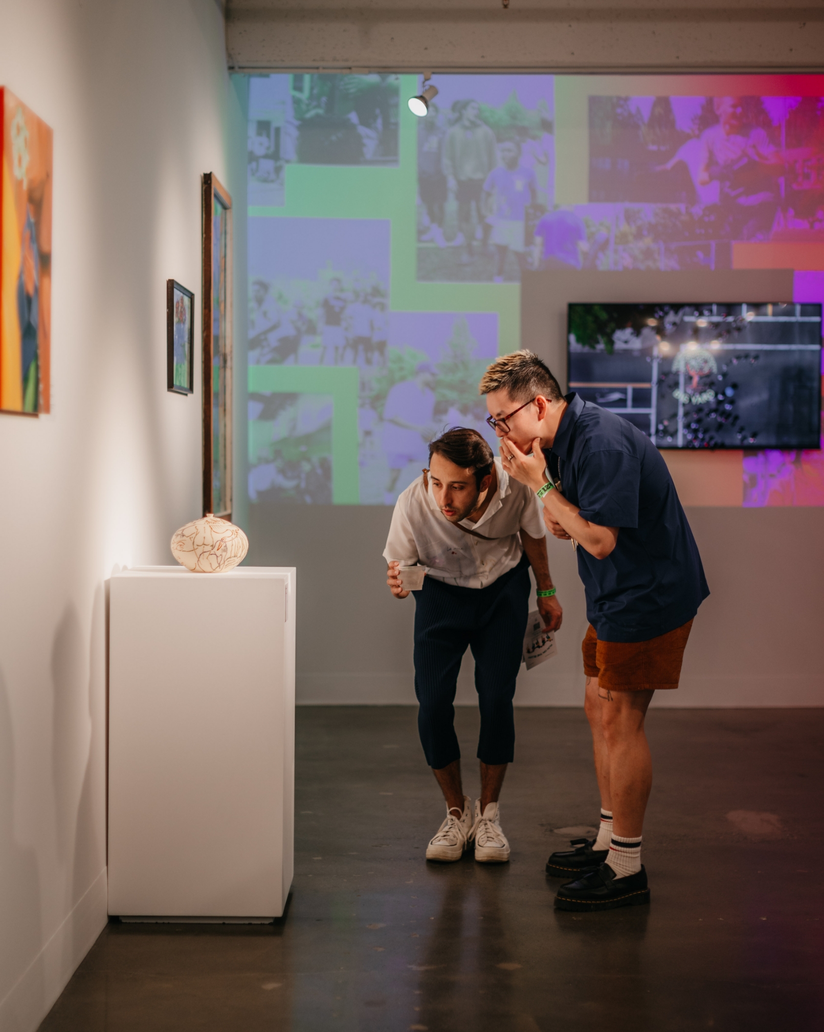 Two people closely examining an object displayed on a pedestal in a contemporary art gallery with various artworks on the walls and a projected collage in the background.