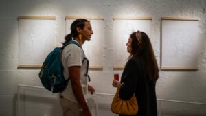 Two people stand in an art gallery discussing framed sketches on a white wall. One holds a drink and the other wears a backpack.
