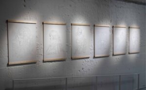 Six framed sketches are displayed on a white textured wall in an art gallery, each sketch subtly lit from above.