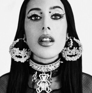 Black and white photograph of a person with long dark hair, wearing large hoop earrings, a choker with studs, and heart-shaped makeup above their eyes.