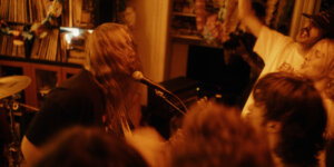 A band performs energetically in a dimly lit room filled with an enthusiastic audience. Records are visible in the background.