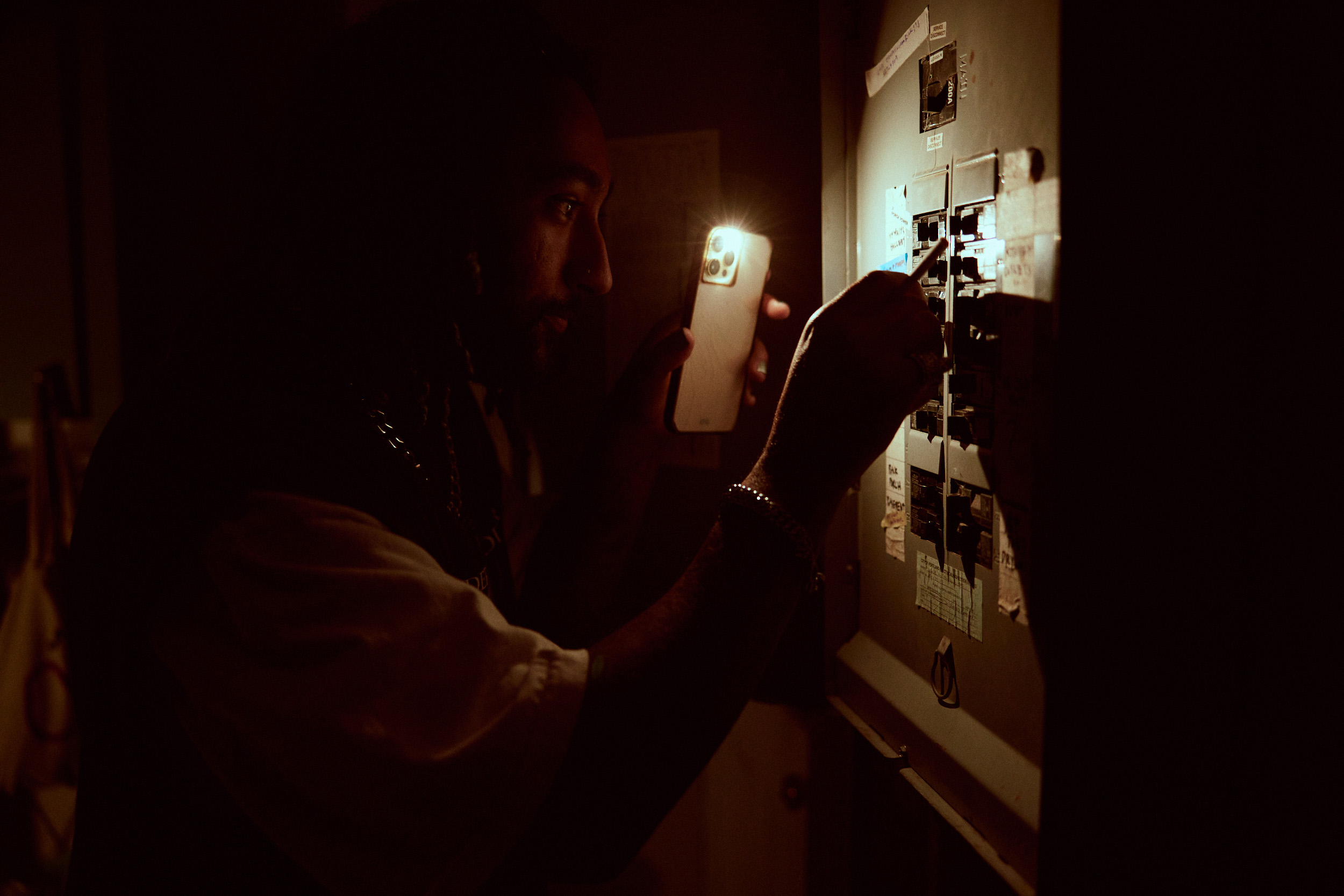Person using a smartphone flashlight to inspect a circuit breaker panel in a dimly lit room.