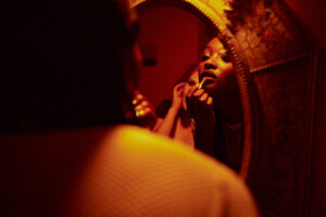 A person applies lipstick while looking into an ornate, round mirror in a dimly lit room.