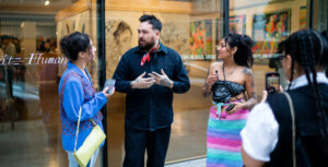 Three people stand in conversation outside an art gallery, holding phones. The gallery displays various paintings on the walls.