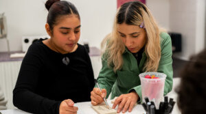 Two people are sitting at a table. One person is drawing on paper, while the other observes. A container of strawberries is on the table.
