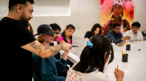 A group of people participate in an art class. The instructor, with tattoos, points at a student's drawing while others work at a table covered with papers and art supplies.