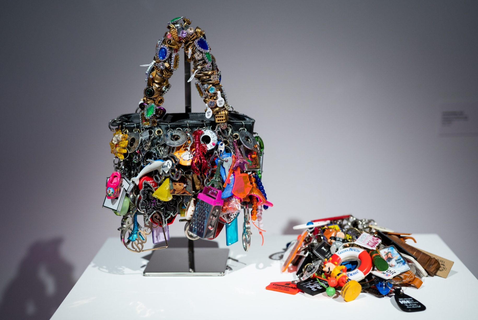 A handbag made of various colorful trinkets is displayed on a stand next to a pile of similar items on a white surface.