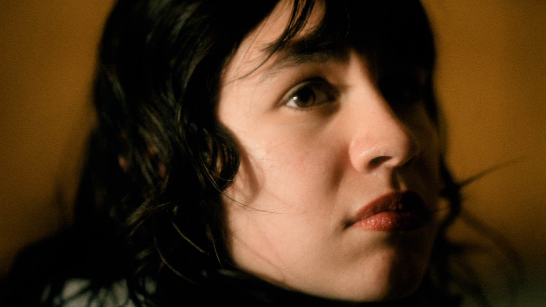 A person with dark hair looks upward against a plain background, with light illuminating one side of their face.