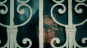 A person wearing glasses looks out from behind a white, decorative metal grill, with their face partially obscured by bars and shadow.