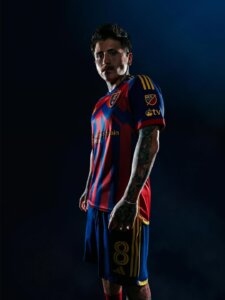A male soccer player in a red and blue Real Salt Lake jersey with number 8 stands against a dark background, looking at the camera.