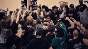 A crowd of people at a concert participates in moshing and crowd surfing; many wear dark shirts and appear energetic and engaged.