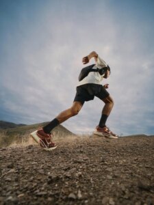 A person wearing athletic gear runs uphill on a rocky trail with cloudy skies and distant hills in the background.