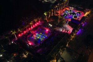 Aerial view of a brightly lit outdoor skate park event at night, surrounded by palm trees and crowds, with colorful lights illuminating the scene.