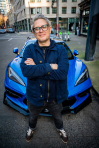 Person with glasses and gray hair stands with crossed arms in front of a blue sports car on a city street.