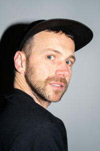 A man with short hair, light eyes, and facial stubble wearing a black cap and shirt looks over his shoulder against a plain background.