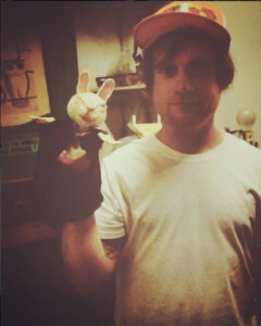 A man wearing a white t-shirt and orange cap holds a hand puppet resembling a rabbit made from a white object, indoors.