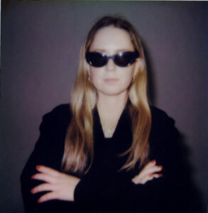 A person with long blond hair wearing dark sunglasses and a black outfit stands with arms crossed against a plain background.