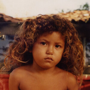 A young child with curly hair looks at the camera with a neutral expression, sitting outdoors with a tiled roof visible in the background.