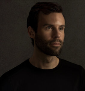 A man with short dark hair and a beard, wearing a black shirt, looks slightly to the side against a dark, plain background.
