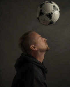 A man in a dark hoodie looks up, balancing a black and white soccer ball on his forehead against a plain dark background.