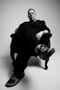 A person sits in a large armchair with legs crossed, wearing dark clothing and sneakers, looking directly at the camera in a plain studio setting.
