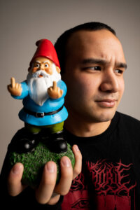 A person holds a garden gnome figurine with both hands raised, showing its middle fingers. The person looks to the side with a neutral expression.
