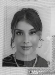 Black and white headshot of a young woman with long hair pulled back, wearing hoop earrings, a necklace, and a dark top, looking straight at the camera.
