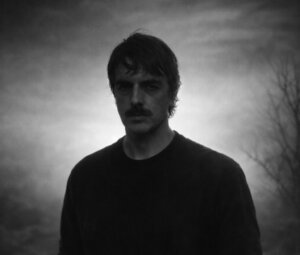 A man with a mustache and dark hair stands outdoors at dusk or dawn, with a bare tree and cloudy sky in the background.