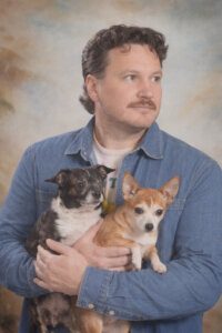 A man in a denim shirt holds two small dogs, one black and white and one tan and white, in front of a painted studio backdrop.