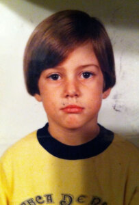 A young child with straight brown hair and a neutral expression wears a yellow shirt with dark trim, facing the camera against a plain background.
