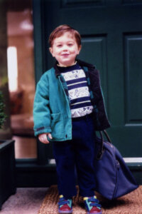 A young child stands in front of a green door, wearing a teal jacket, patterned shirt, navy pants, and colorful shoes, holding a blue bag.