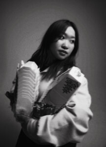 A young woman in a hoodie holds a stack of takeout containers and packaged food, looking to the side in a black and white portrait.