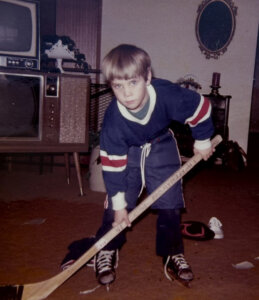 A young boy in hockey gear poses with a hockey stick indoors, standing on carpet in a living room with vintage furniture and a TV in the background.